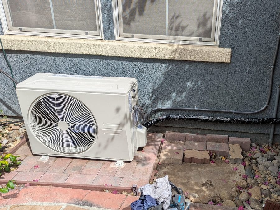 Small ductless-style outdoor unit on a brick paver pad beside a stucco wall with an insulated refrigerant line, Gilroy, CA